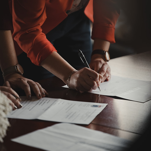 Close-up view of a person wearing a red blazer holding a pen and signing a paper document on a wooden desk alongside other business documents.