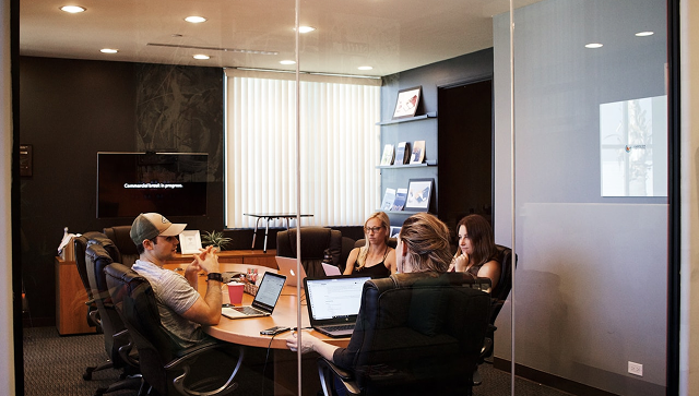 A group of advisors sitting around an oval conference table with laptops during a collaborative strategy meeting inside a glass-walled office.
