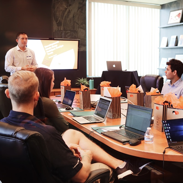 A presenter standing in front of a digital screen giving a presentation to colleagues seated around a conference table with open laptops and gift bags.