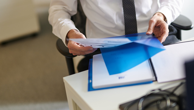 Close-up view of a professional dressed in a white dress shirt and tie sitting at a desk, opening a blue plastic folder to review printed business documents.