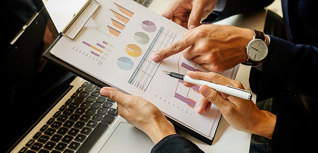 Two business professionals analyzing financial performance report charts on a clipboard next to an open laptop, with one person pointing to data and another holding a fountain pen.