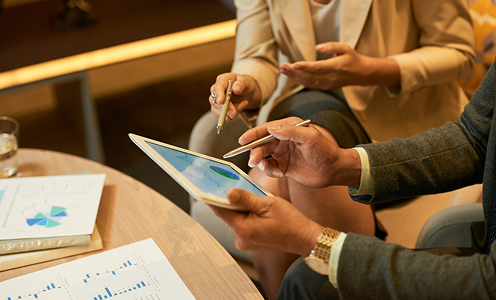 A close-up view of two business professionals holding pens and analyzing charts on a digital tablet at a coffee table with financial reports.