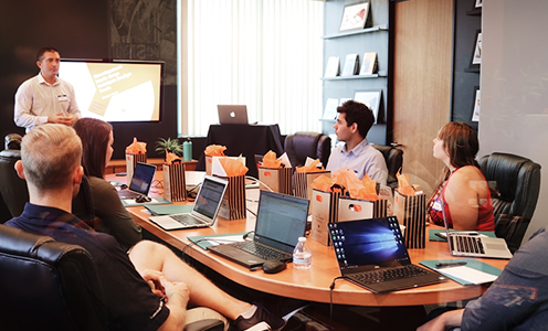 A presenter standing in front of a digital screen giving a presentation to a team seated around a conference table with open laptops.
